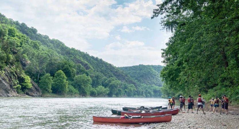 A group of outward bound students stand in a circle on the shore of a river with their canoes nearby. The river is lined by wooded hills. 